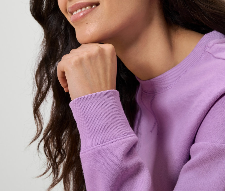 Horní část těla ženy s tmavými vlnitými vlasy, která se opírá rukou o bradu. Má na sobě světle fialovou mikinu.