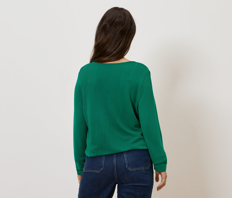 Zena stojí zády a má na sobě zelený svetr a džíny.