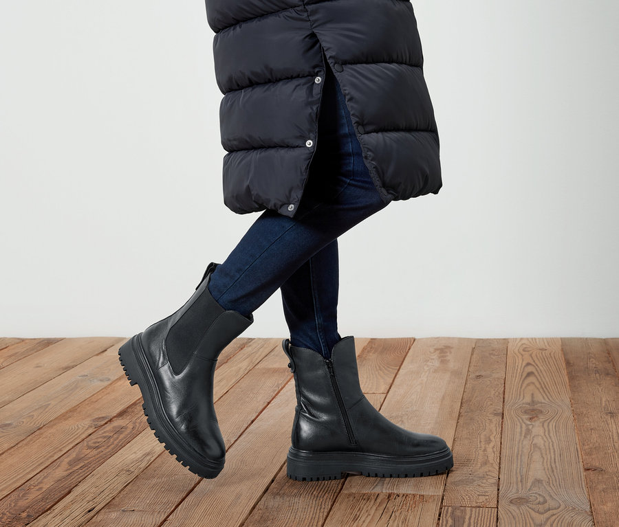 Nohy osoby v džínových kalhotách a černých kožených botách na dřevěné podlaze. Osoba má na sobě dlouhou černou bundu.