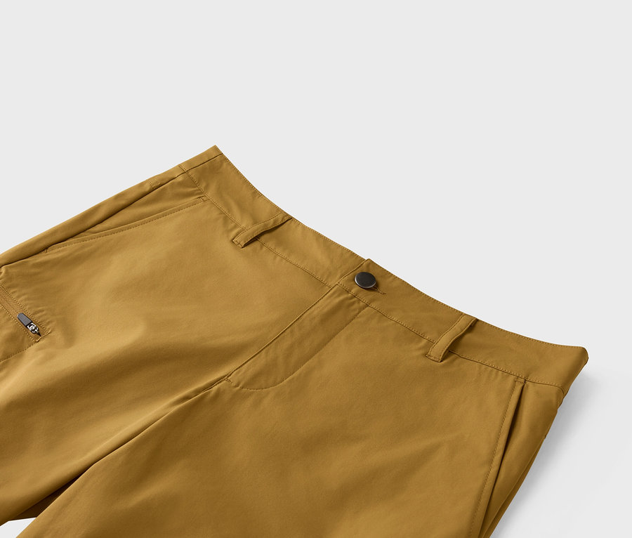 Detailní záběr na okrové kalhoty se zapínáním na knoflík a kapsou na zip.