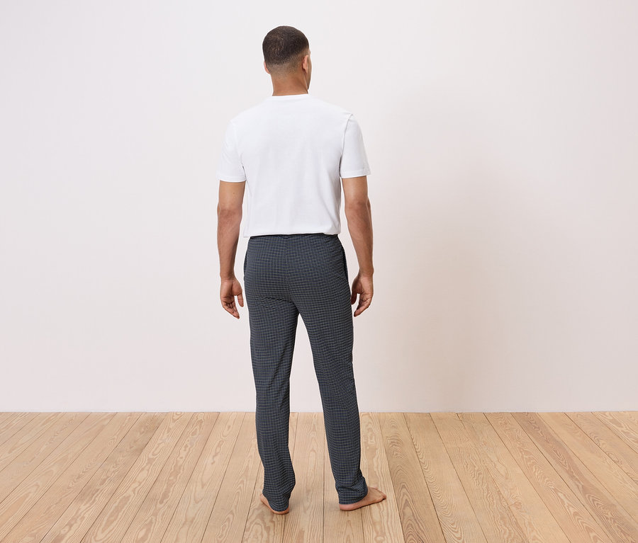 Muž zády k fotoaparátu stojí na dřevěné podlaze a má na sobě bílé tričko a tmavé kostkované kalhoty.