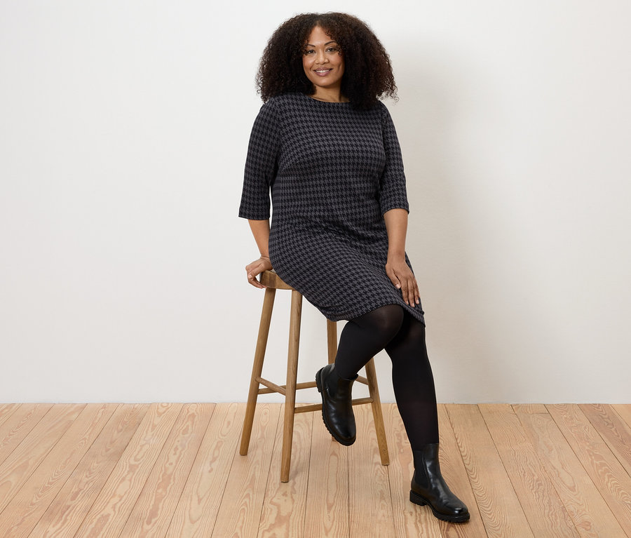 Žena sedí na stoličce a má na sobě žerzejové šaty s žakárovým vzorem kohoutí stopy.