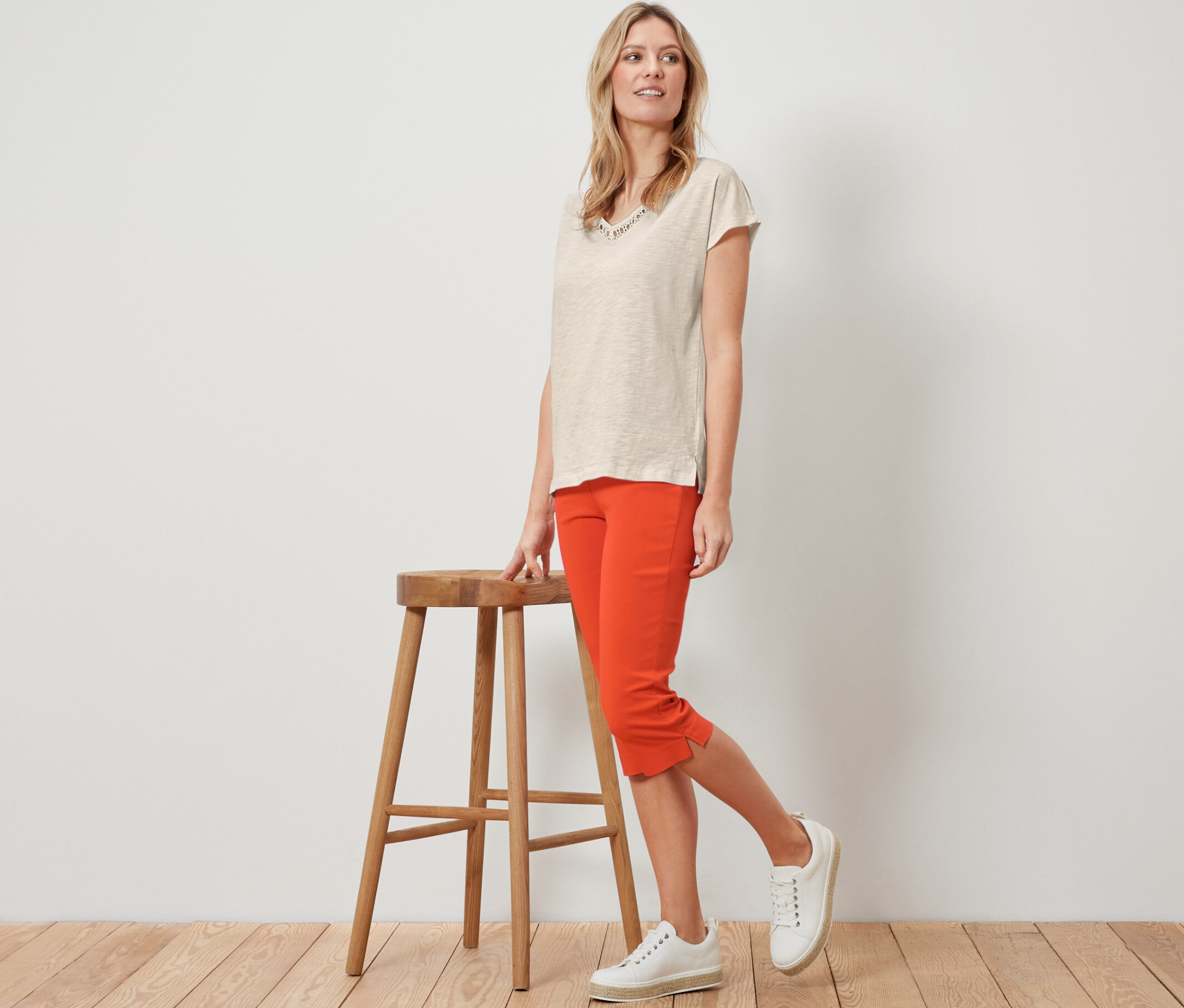 Blondýna pózuje vedle dřevěné stoličky v oranžových 3/4 strečových kalhotách.