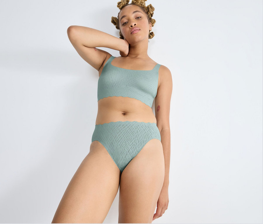 Žena s blond dredy stojí u bílé zdi, oblečená ve vysoce vykrojených kalhotkách sloggi ZERO Feel Bliss High leg v barvě misty turquoise a sladěném topu. Jednu ruku má zvednutou za hlavou a dívá se do kamery. Oba kousky mají jemný vzor a vlnité okraje.