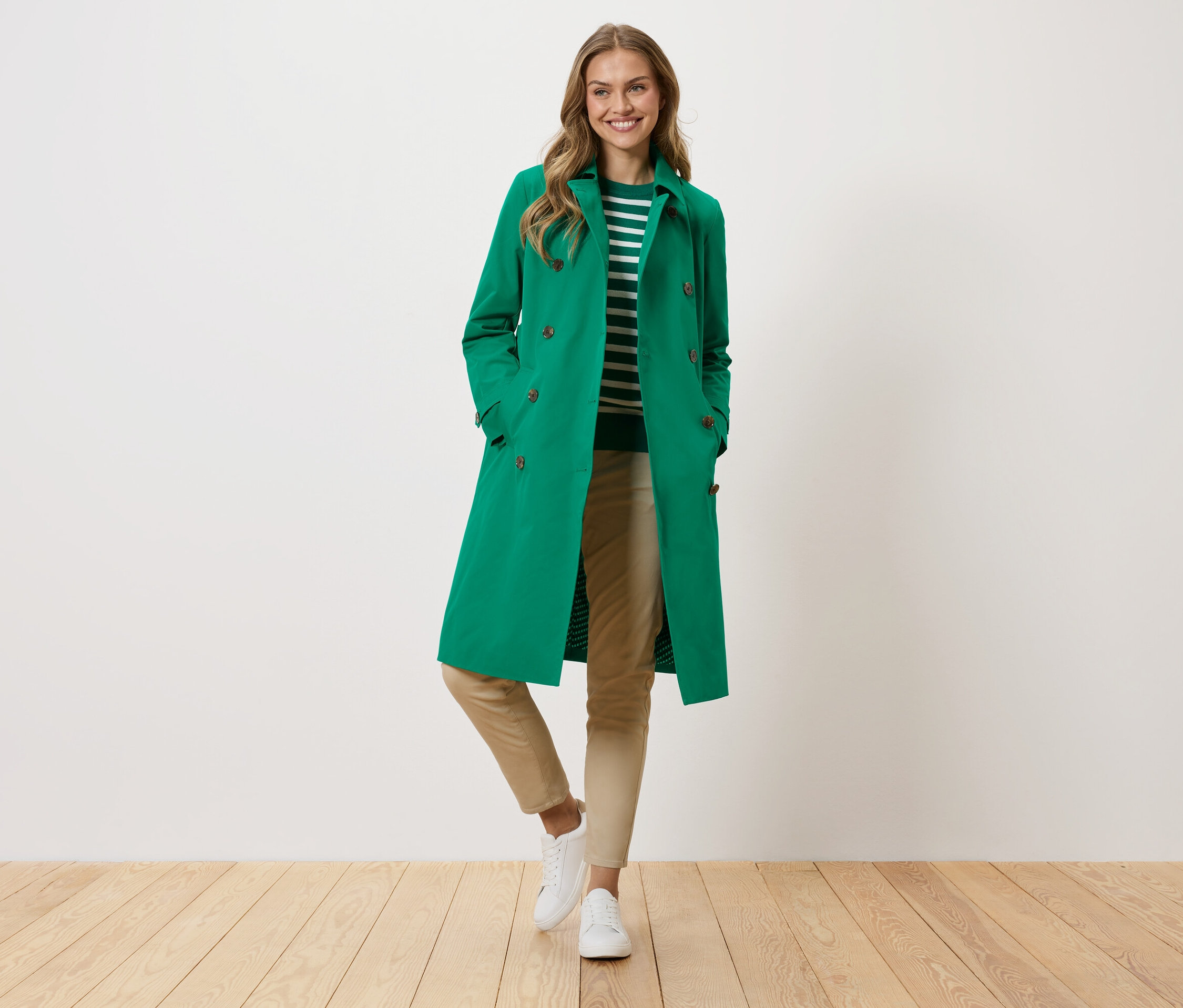 Žena v zeleném kabátě, pruhovaném svetru, béžových kalhotách a bílých teniskách stojí před bílou zdí.