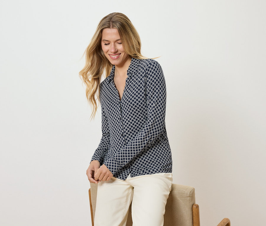 Žena s dlouhými blond vlasy v vzorované blůze a světlých kalhotách sedí na židli a usmívá se.