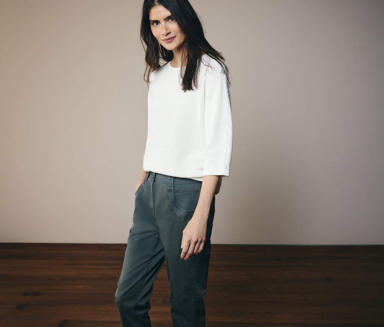 Žena v bílém topu a kalhotách CECIL »Scarlett« v tmavě zelené pózuje na světlém pozadí.