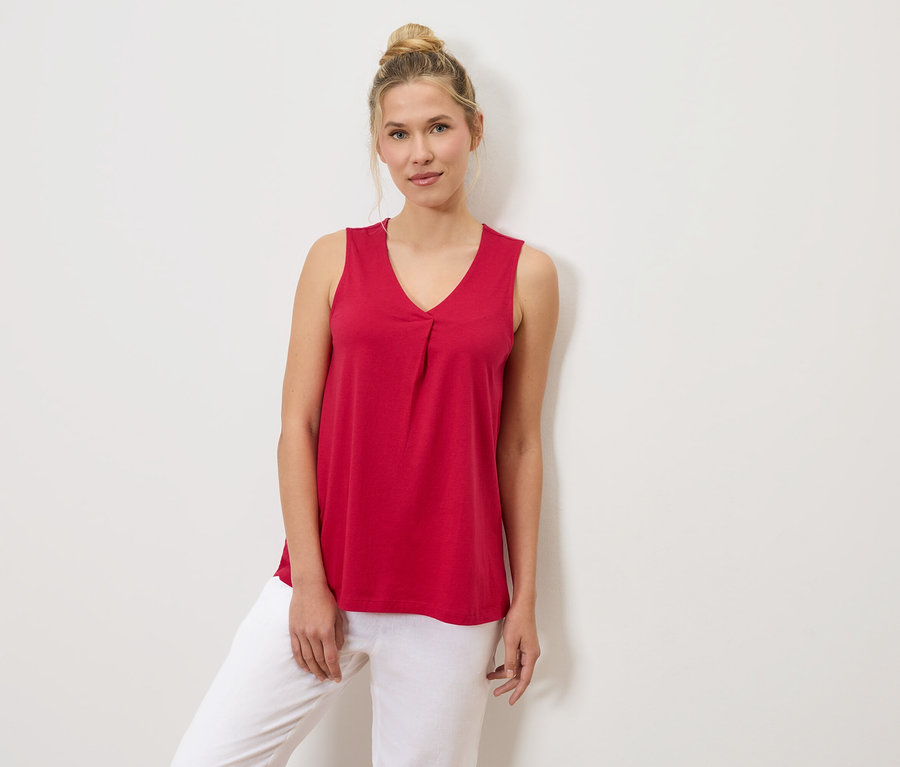 Žena s drdolem má na sobě červený tílko a bílé kalhoty, opírajíc se o bílou stěnu.