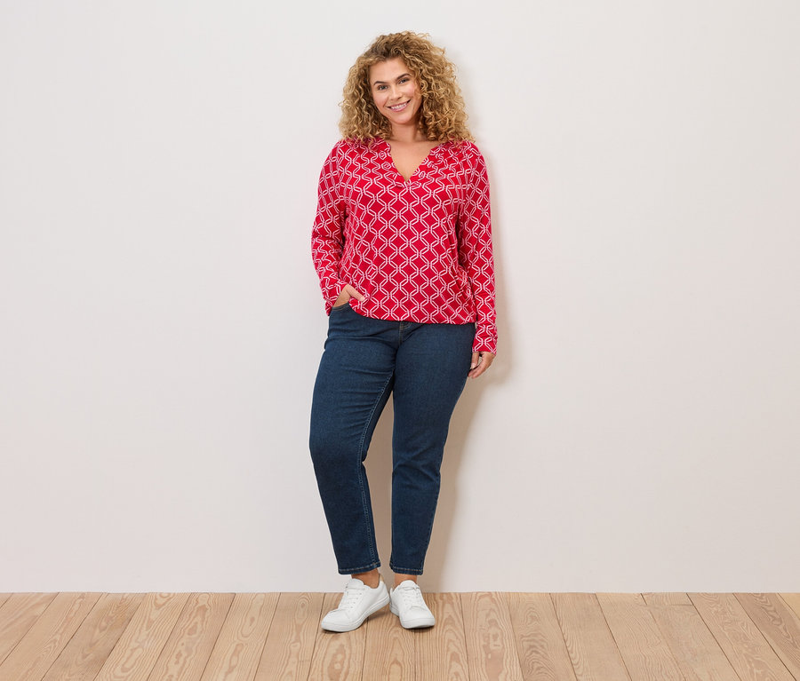 Žena s kudrnatými blond vlasy, v červené halence s bílým vzorem, džínách a bílých teniskách stojí před zdí.