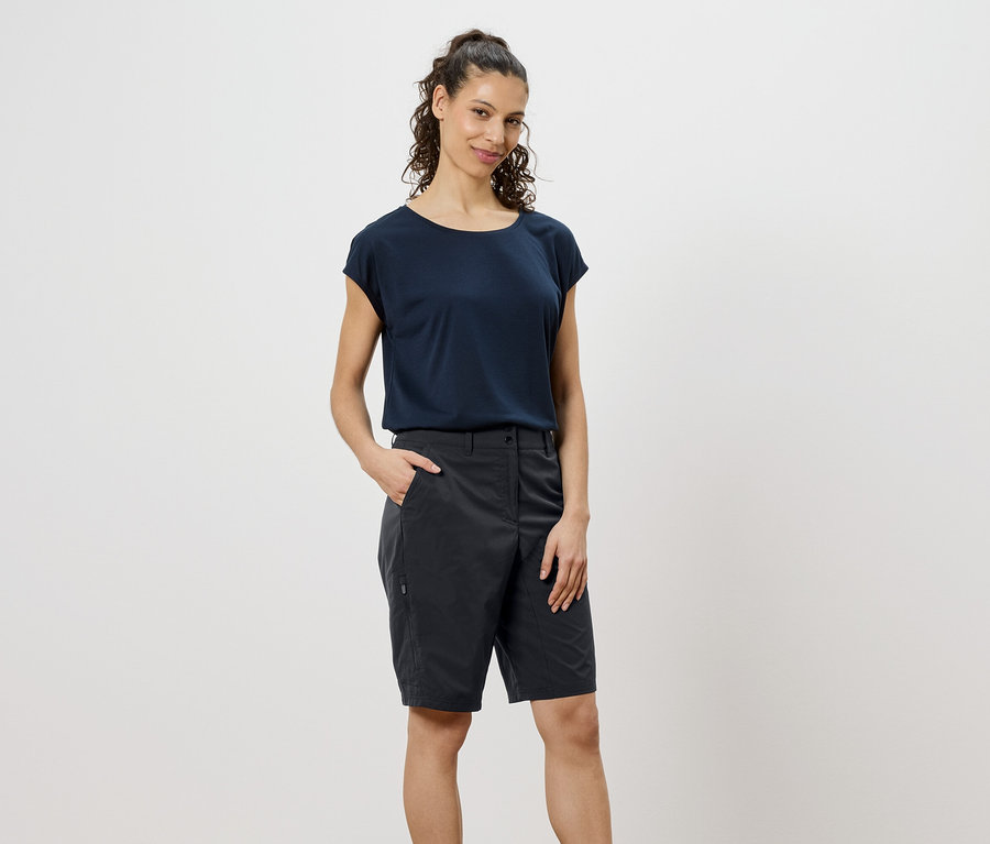 Žena s kudrnatými hnědými vlasy má na sobě tmavě modré tričko a šedé šortky a dává ruku do kapsy.