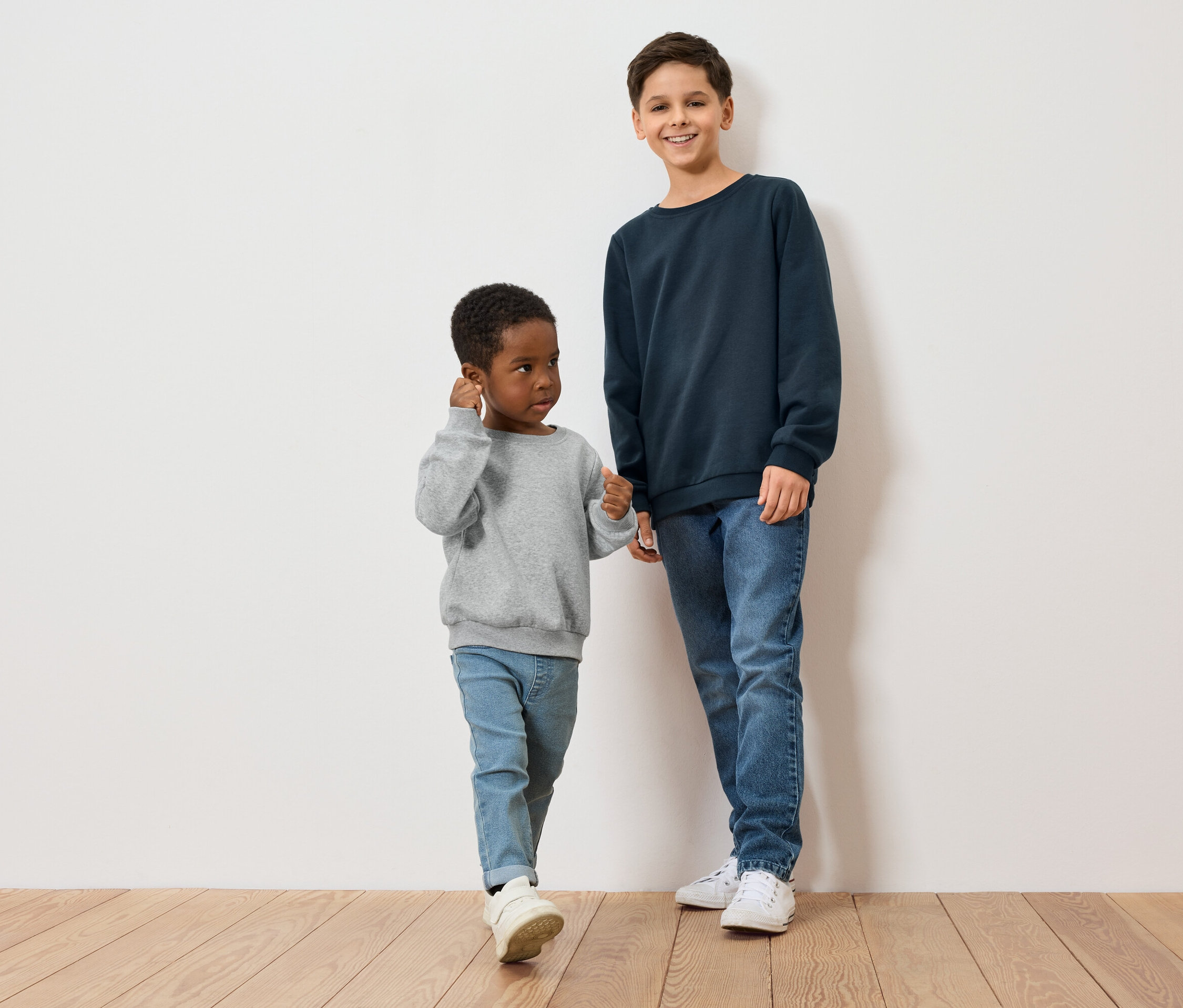 Dva chlapci, jeden s tmavou pletí v šedém svetru, druhý se světlou pletí v modrém svetru, stojí na dřevěné podlaze před bílou zdí.