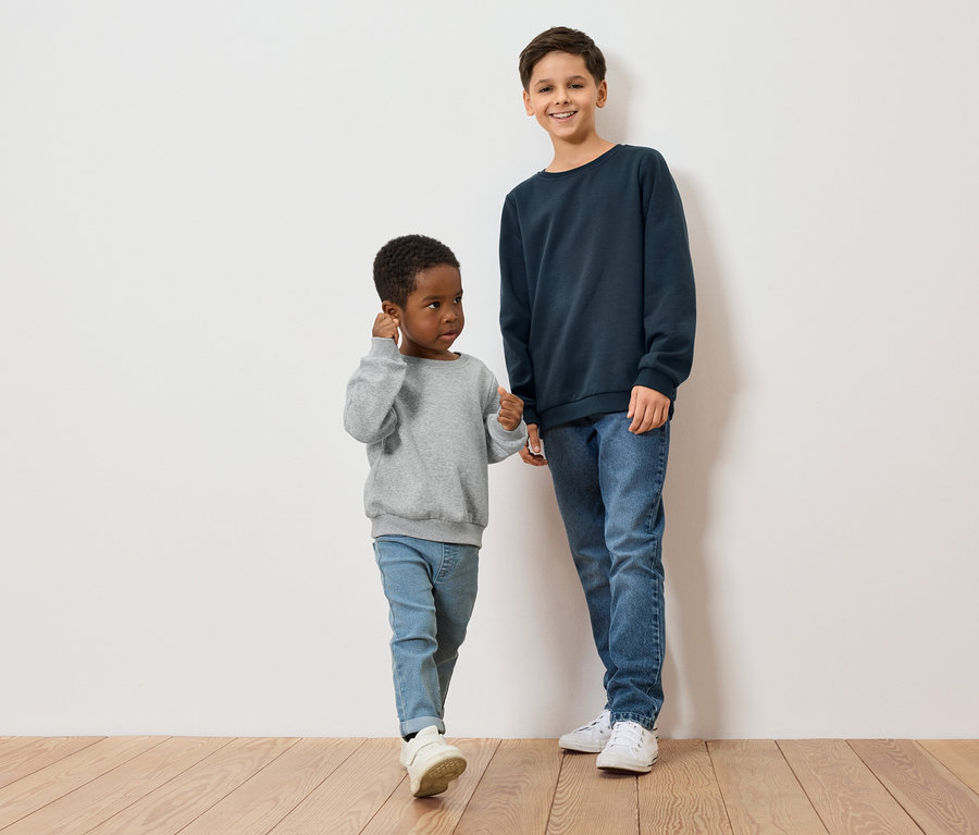 Dva chlapci, jeden s tmavou pletí v šedém svetru, druhý se světlou pletí v modrém svetru, stojí na dřevěné podlaze před bílou zdí.