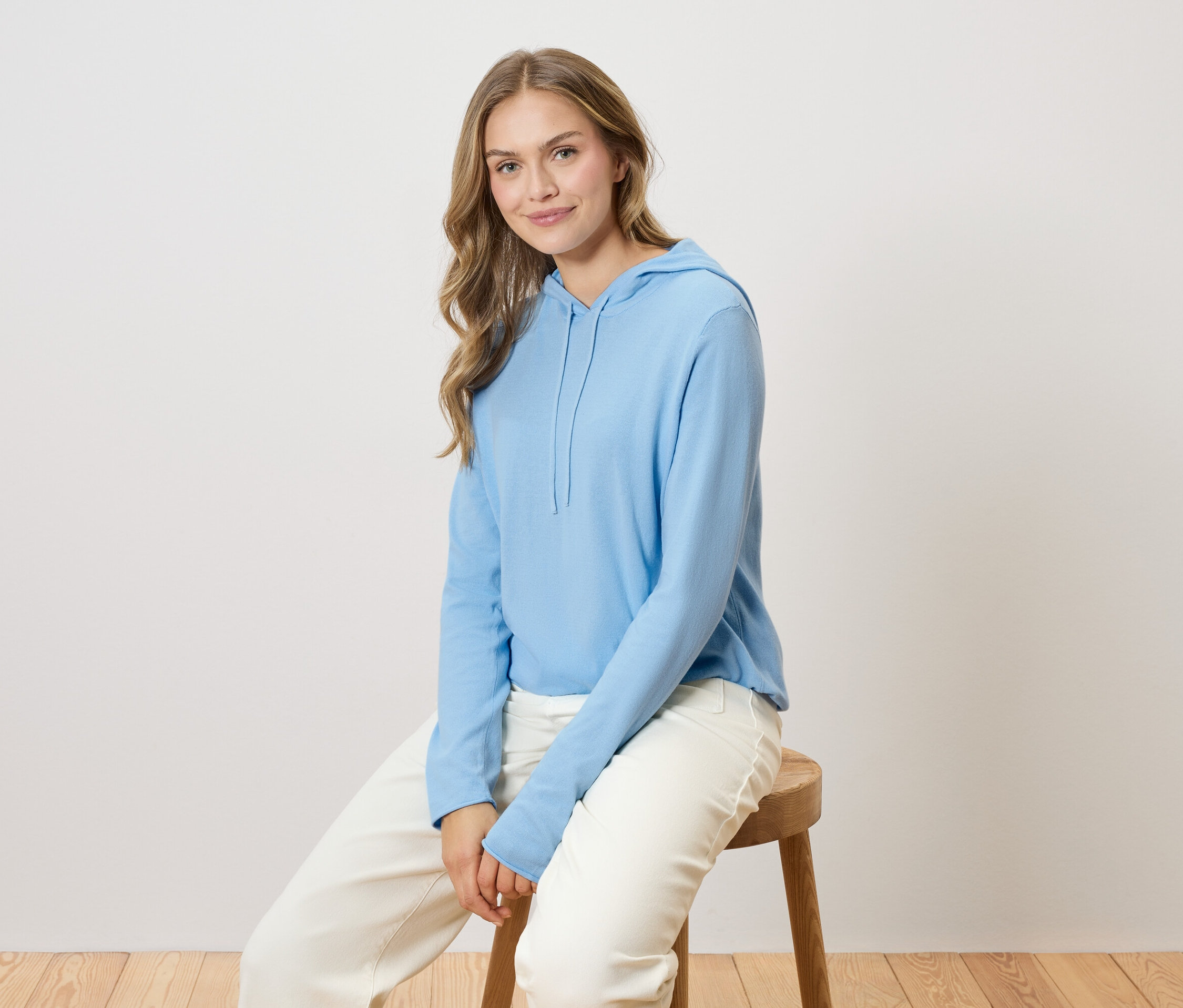 Žena s dlouhými blond vlasy sedí na dřevěné židli a má na sobě světle modrou mikinu s kapucí a bílé kalhoty.