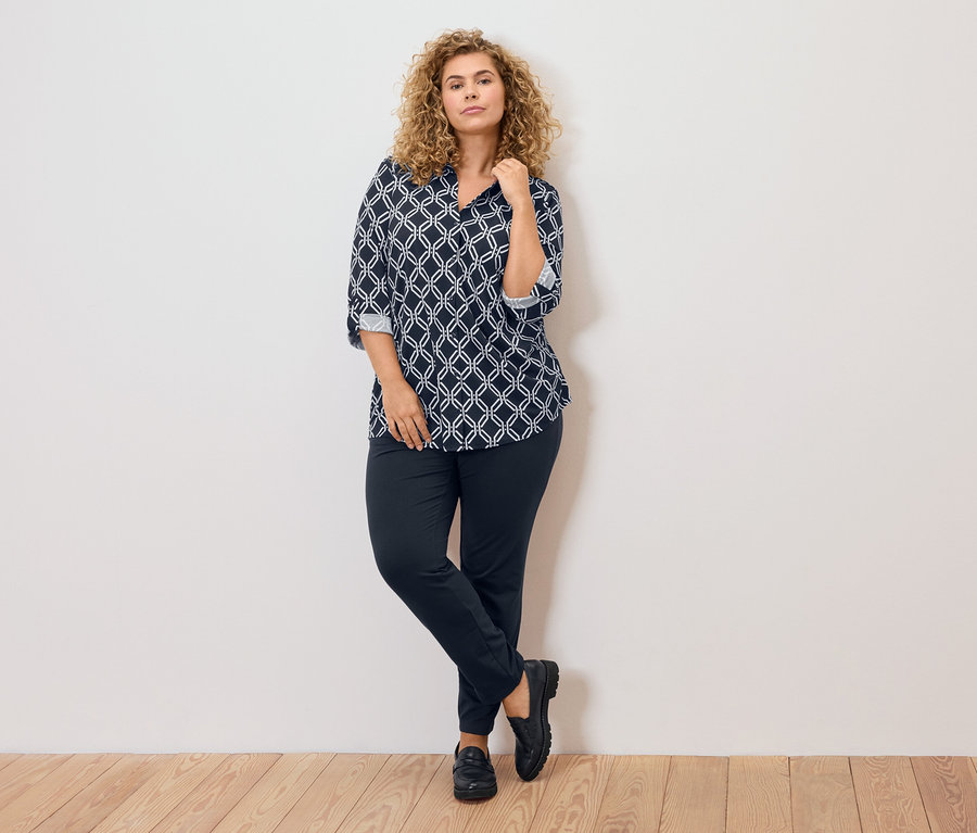 Žena s kudrnatými blond vlasy, oblečená v modro-bílé vzorované halence, tmavé kalhoty a černé boty, opírá se o zeď.