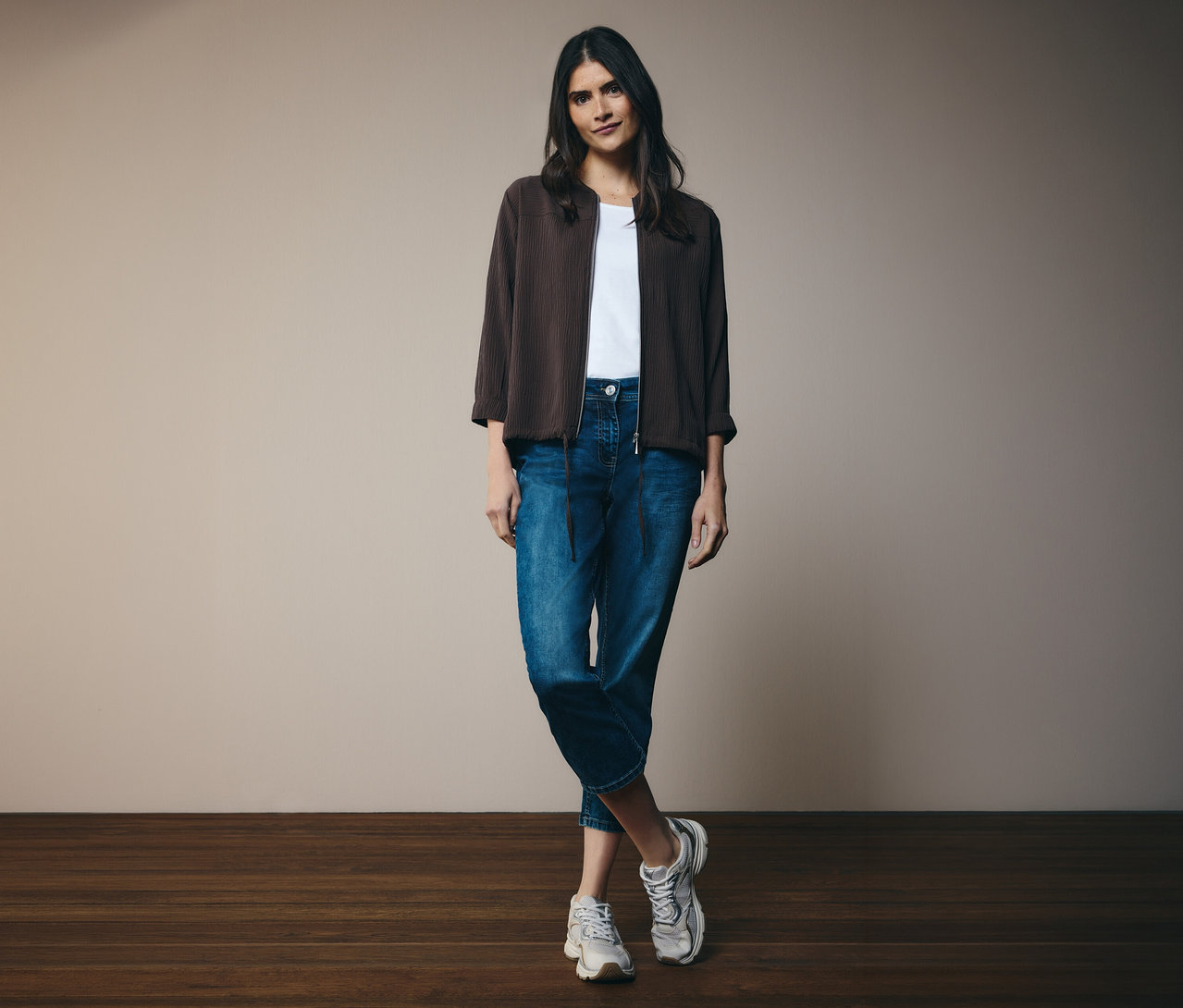 Žena v bílém tričku, džínách a hnědé bundě CECIL. Stojí před zdí.