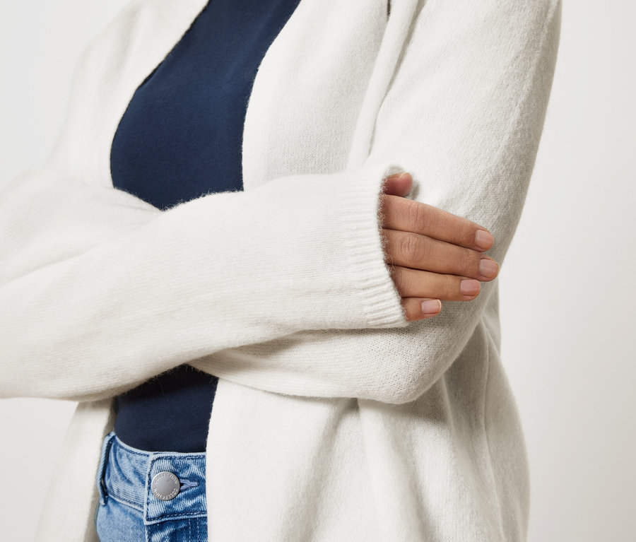 Žena se zkříženýma rukama má na sobě dlouhý bílý kardigan přes tmavě modrý top a modré džíny.