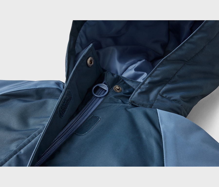 Detail modré bundy ukazující částečně rozepnutý zip, kapuci, patentky a suché zipy na klopě proti větru.