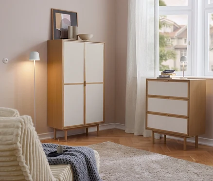 Světlý obývací pokoj s dřevěnou skříní Cabinetschrank se 2 dveřmi a komodou se 3 zásuvkami se světlými čely. Na komodě stojí LED dobíjecí stolní lampa Eglo »Mannera«. Vlevo pohovka, vpravo okno se záclonami.