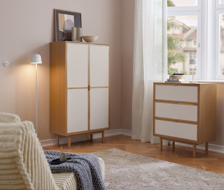 Světlý obývací pokoj s dřevěnou skříní Cabinetschrank se 2 dveřmi a komodou se 3 zásuvkami se světlými čely. Na komodě stojí LED dobíjecí stolní lampa Eglo »Mannera«. Vlevo pohovka, vpravo okno se záclonami.