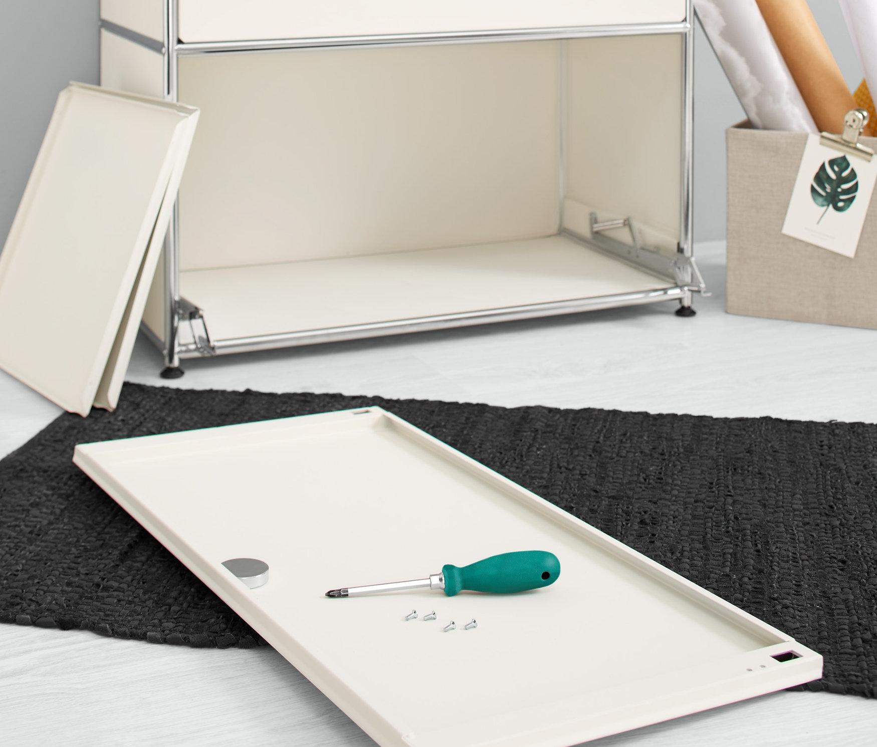 Na podlaze leží bílá doplňková sada kovových výklopných dvířek CN3 s šroubovákem a šrouby vedle komody.