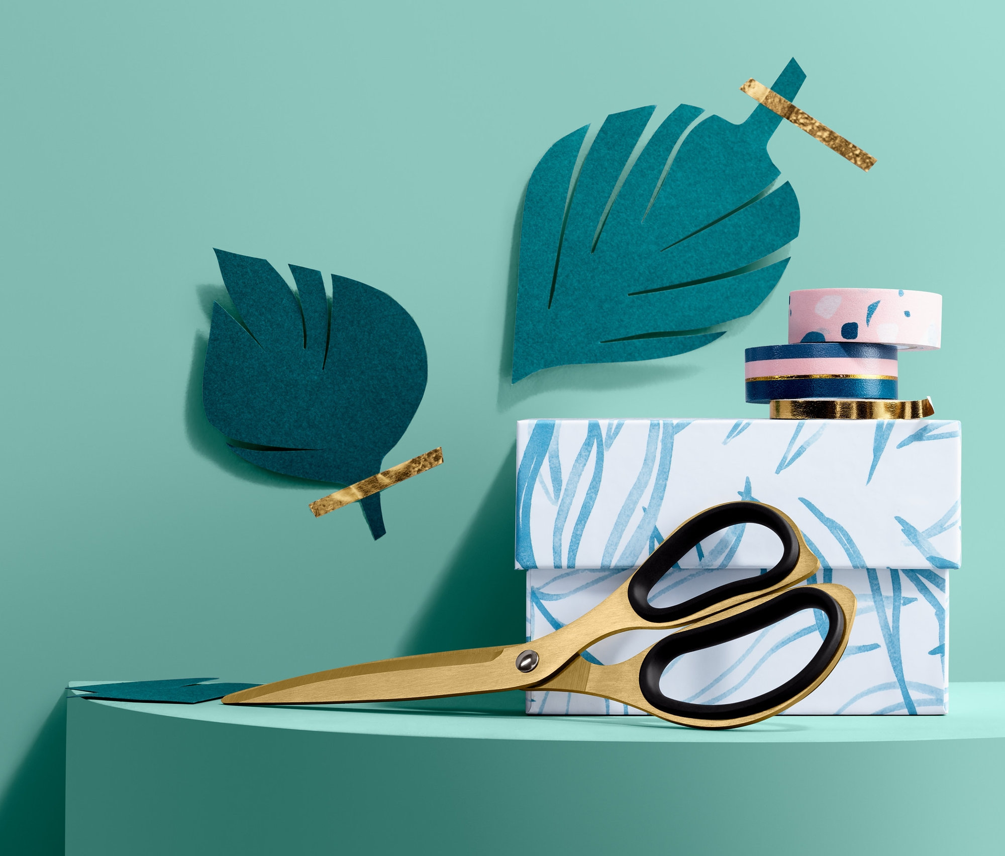 Zlaté kancelářské nůžky leží na krabici s modrým potiskem listů vedle papírových listů.