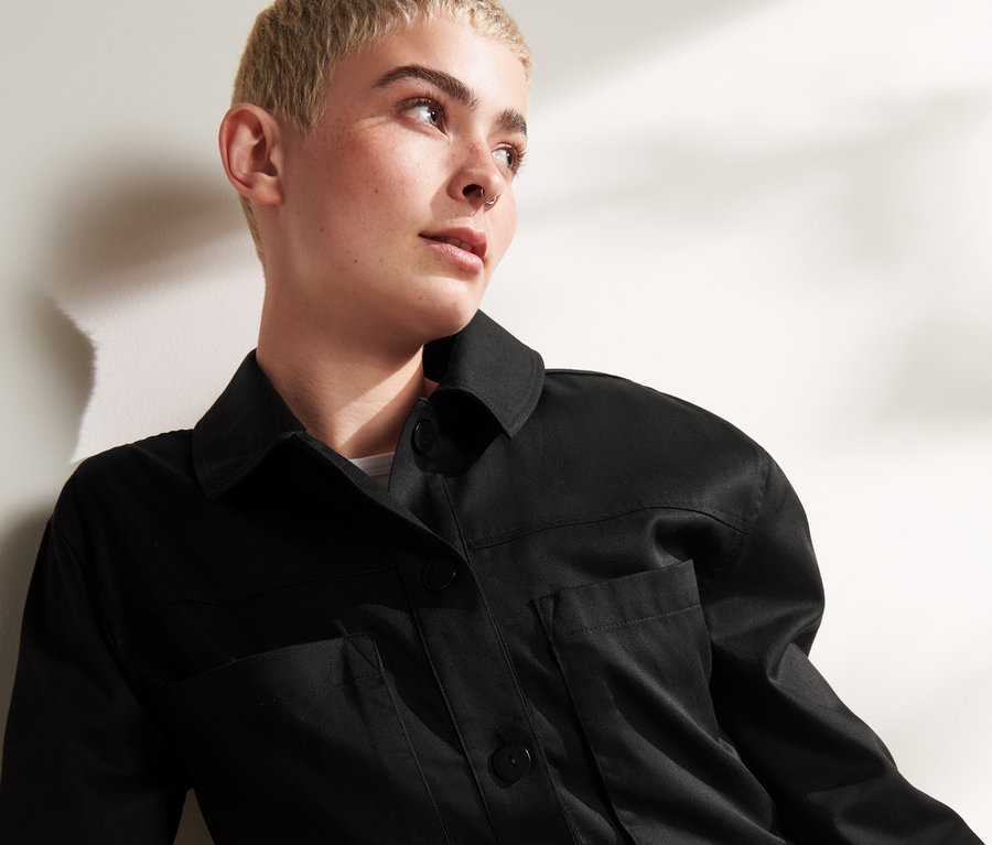 Žena s krátkými vlasy pózuje v černé košilové bundě.