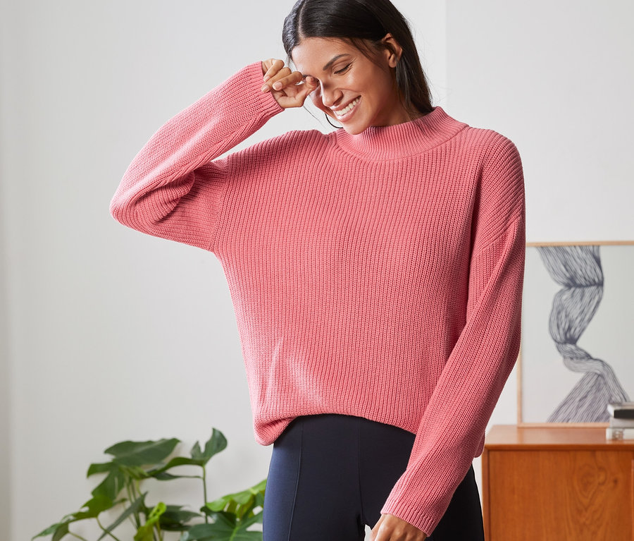 Žena se usmívá a zakrývá si oko rukou, má na sobě pletený svetr se stojáčkem.