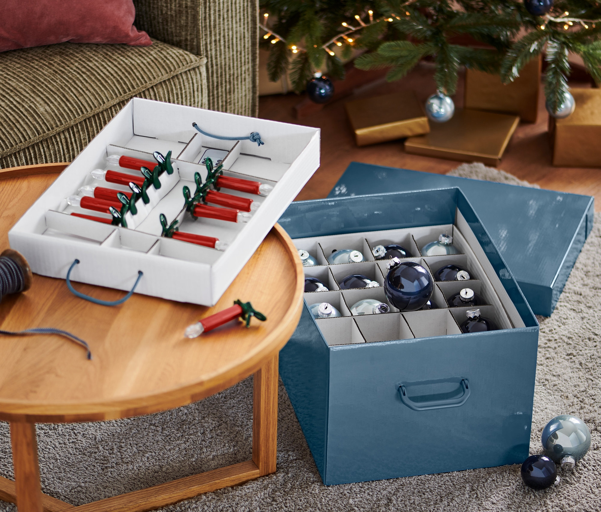 Otevřený úložný box na vánoční ozdoby s červenými svíčkami a modrými ozdobami vedle vánočního stromku.