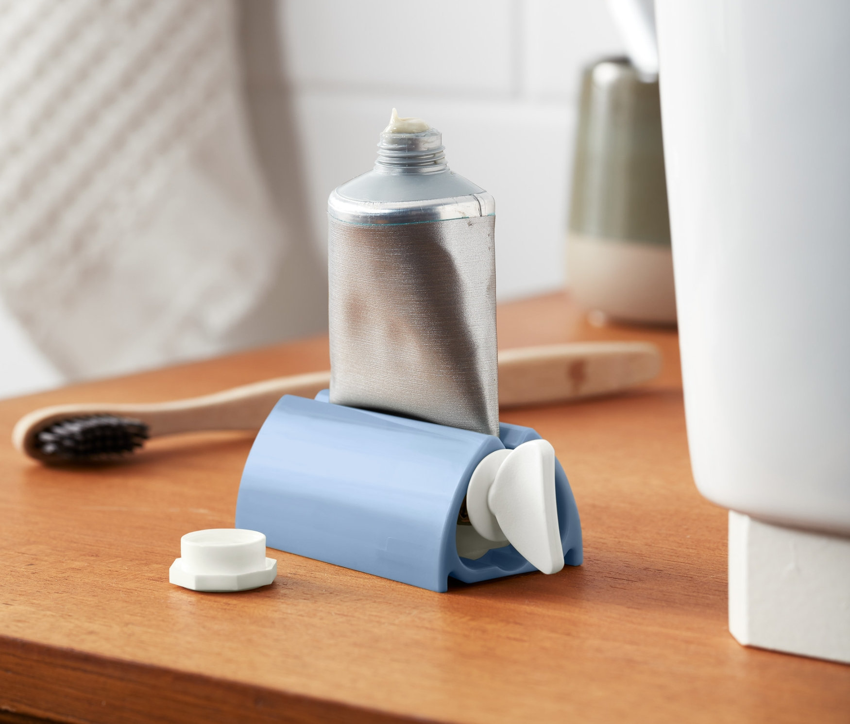 Stříbrná tuba v modrém vytlačovači tub a pasty na dřevěné polici.