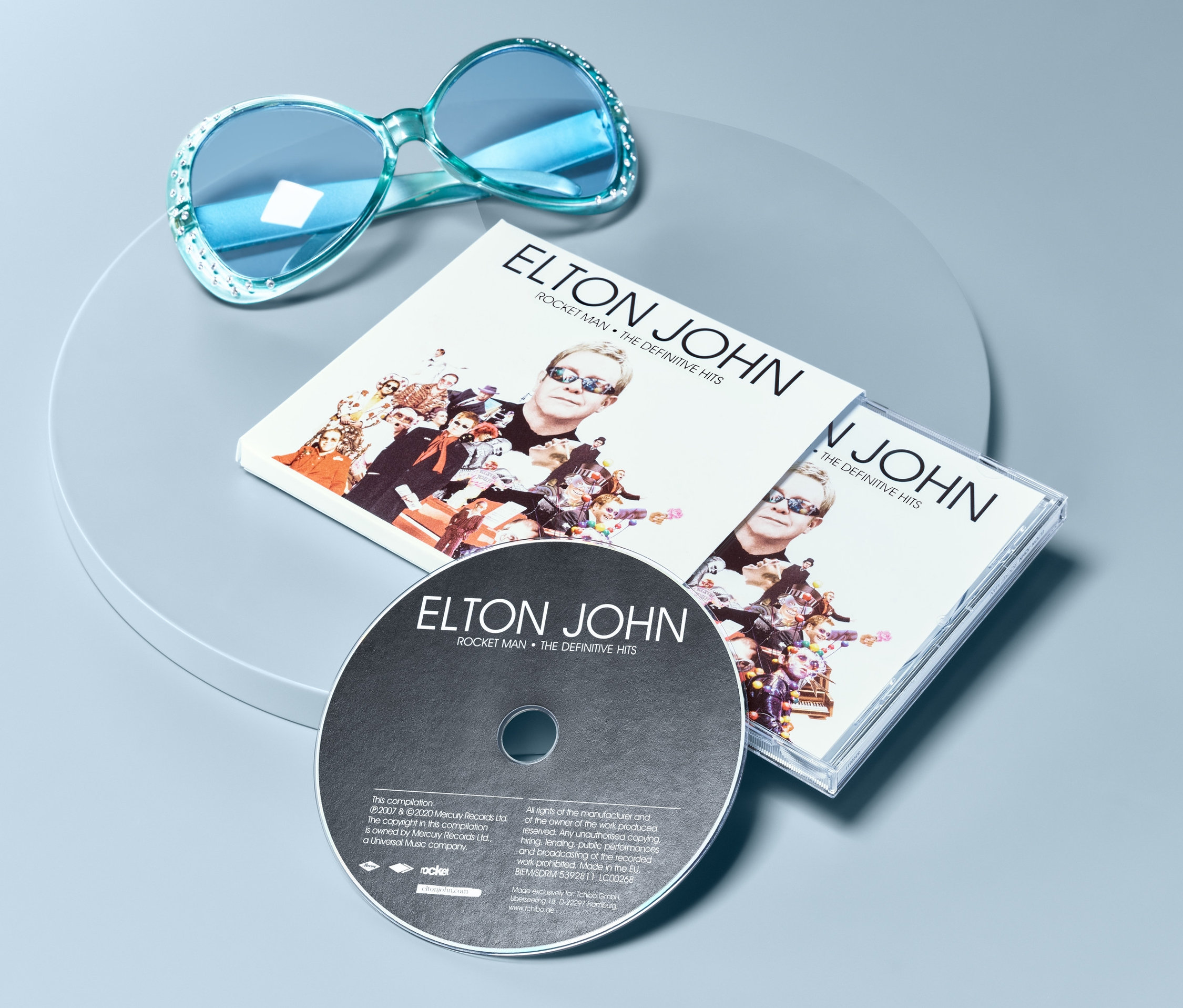 Modré sluneční brýle a CD Elton John – Rocket Man: The Definitive Hits na šedém povrchu.