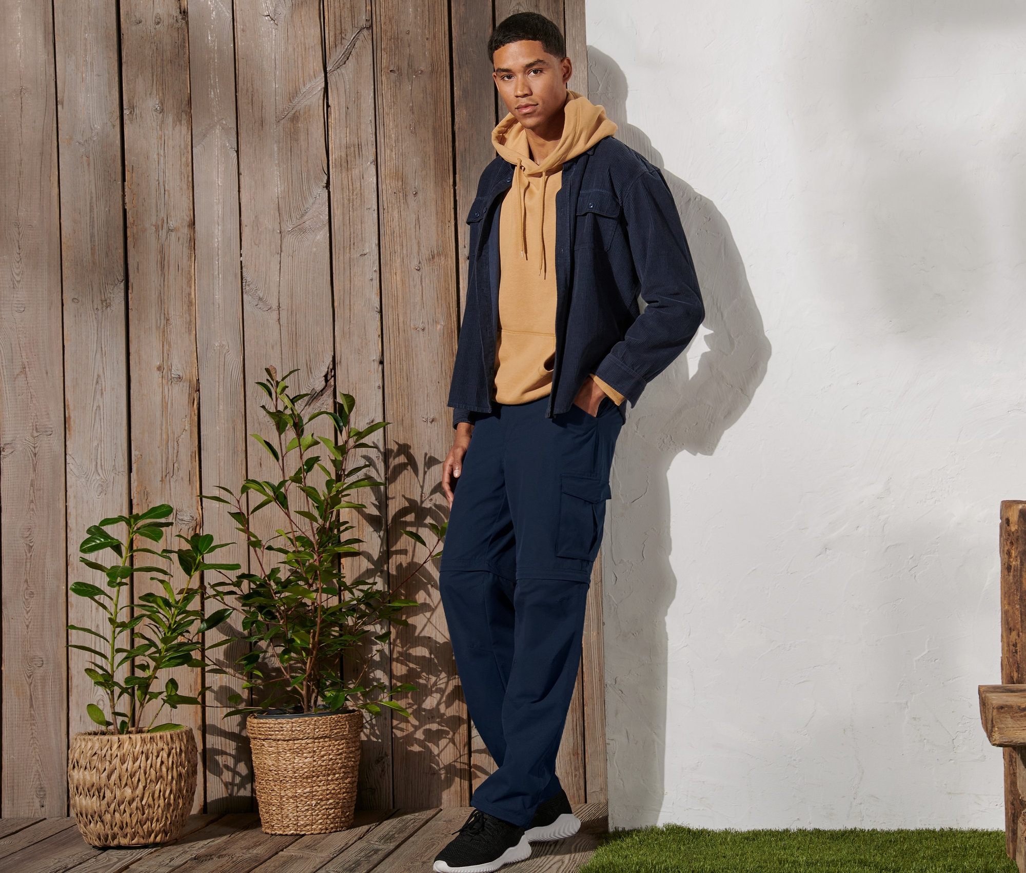 Muž stojí a má na sobě tmavě modré funkční kalhoty 2 v 1, žlutou mikinu a bundu.