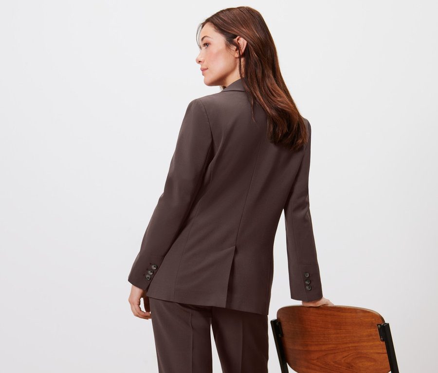 Žena pózuje v tkaném blejzru a kalhotách u dřevěné židle.