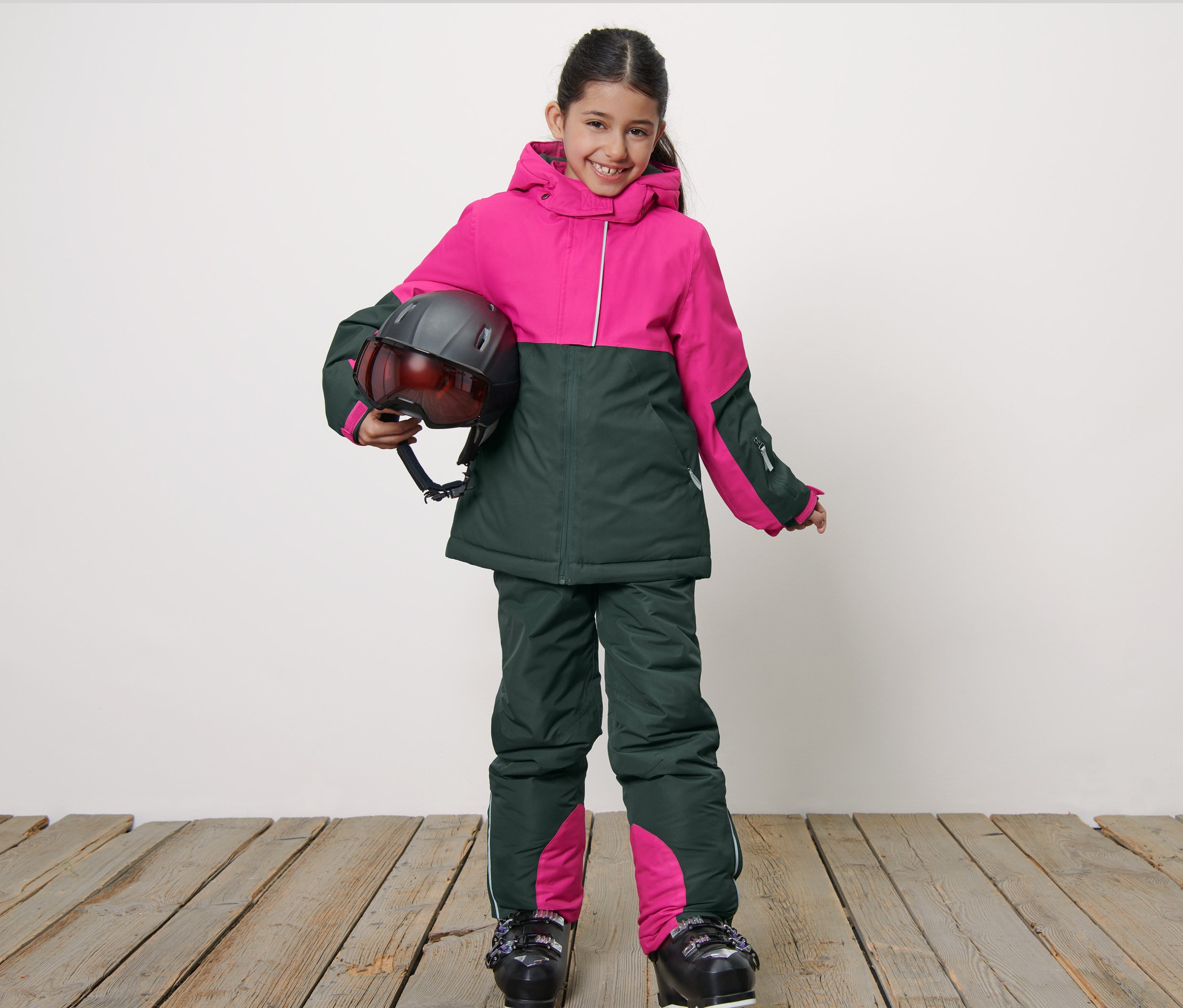 Usmívající se dívka v dětské lyžařské bundě drží helmu.