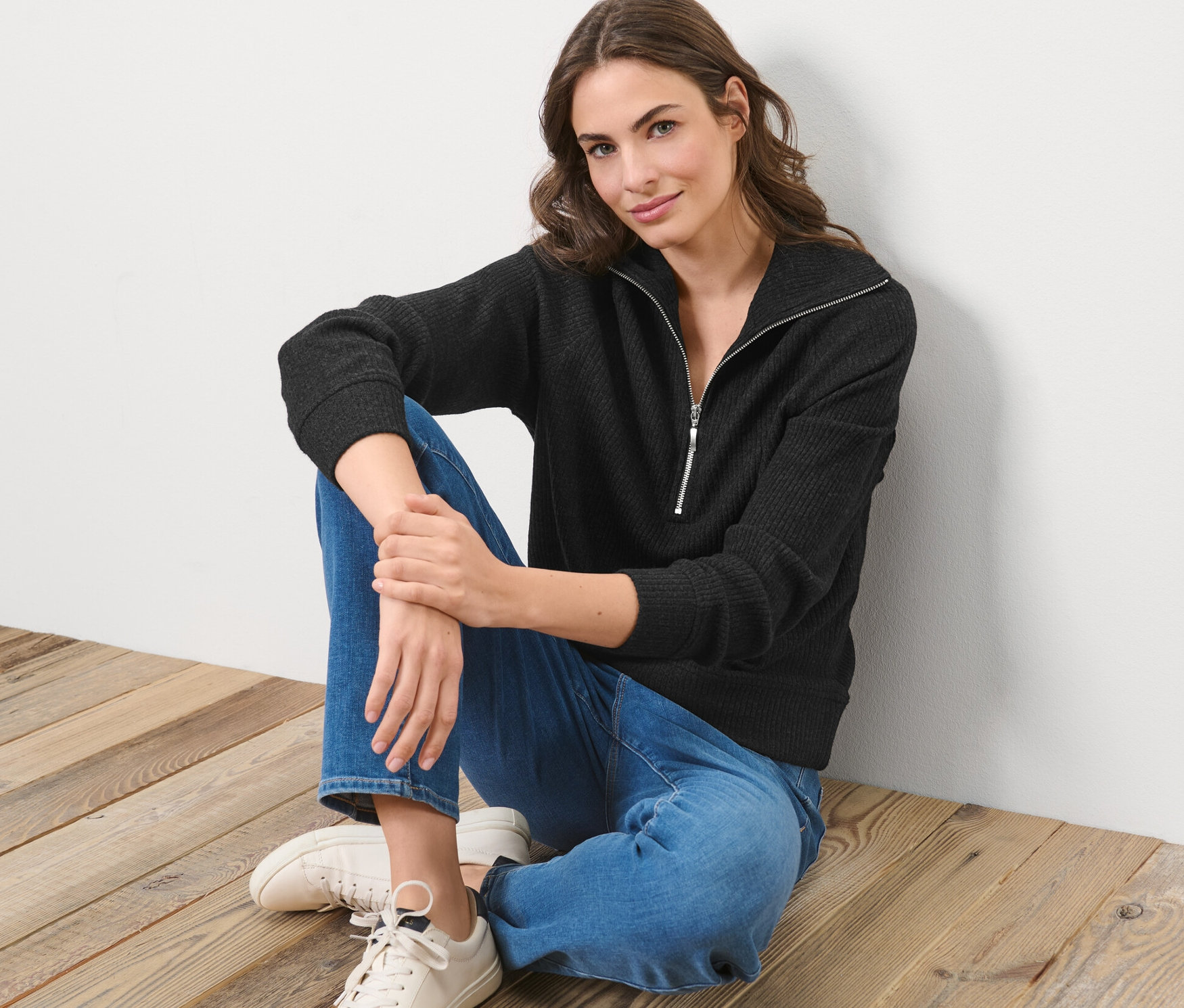 Žena sedí na dřevěné podlaze a má na sobě černou mikinu troyer a džíny.