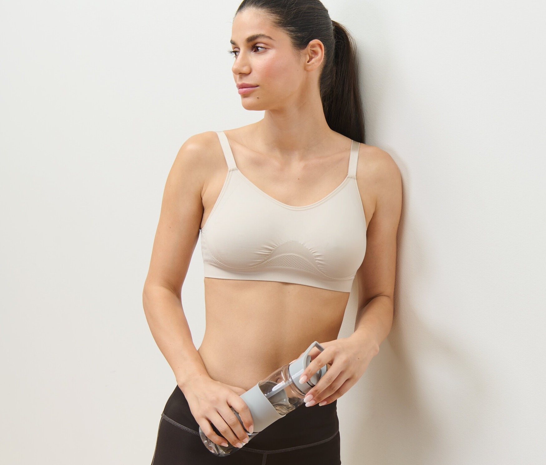 Žena opřená o stěnu drží v ruce láhev s vodou a má na sobě hnědou sportovní podprsenku z 2dílné sady.