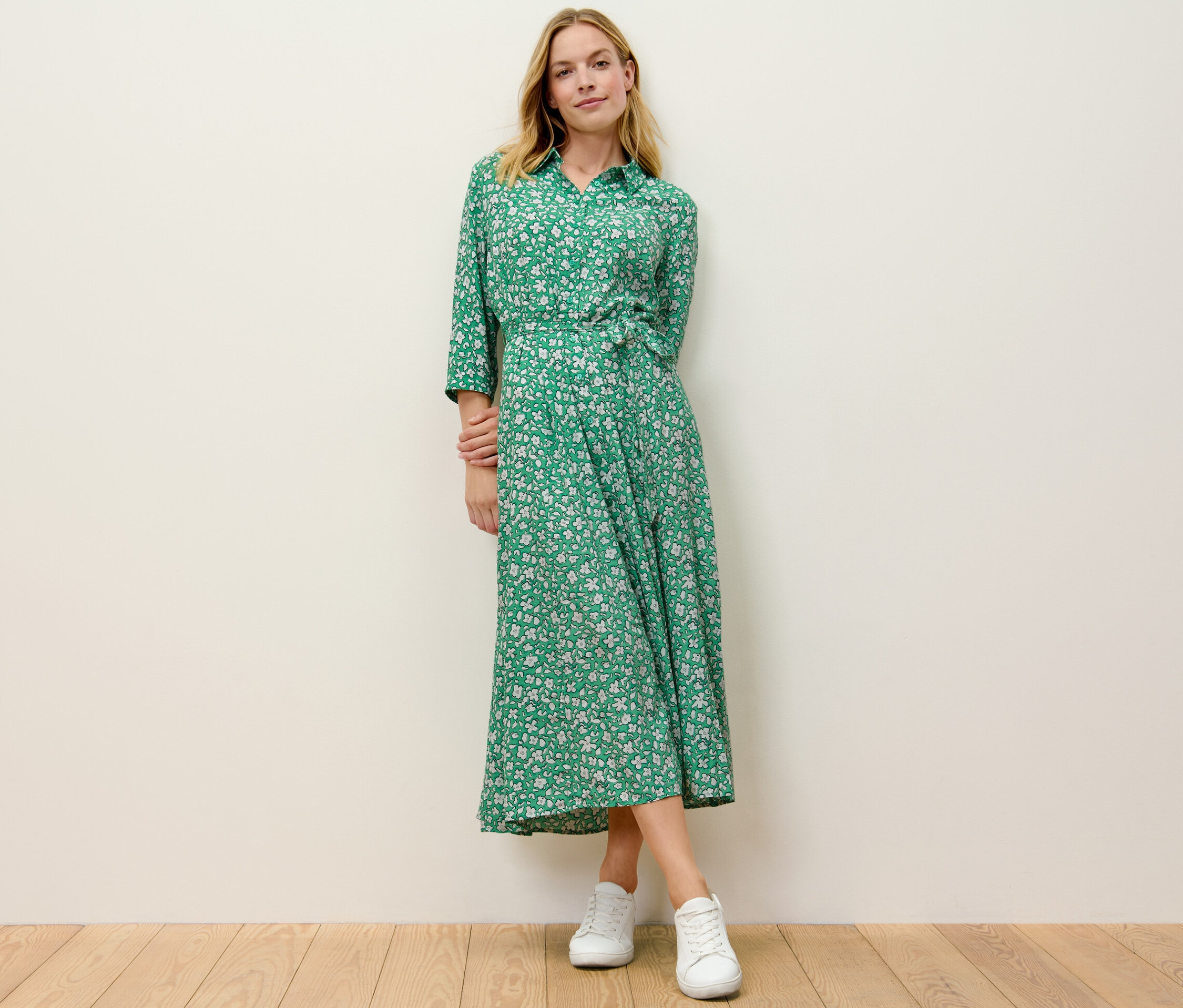 Blondýna stojí a má na sobě zelené košilové šaty s potiskem.