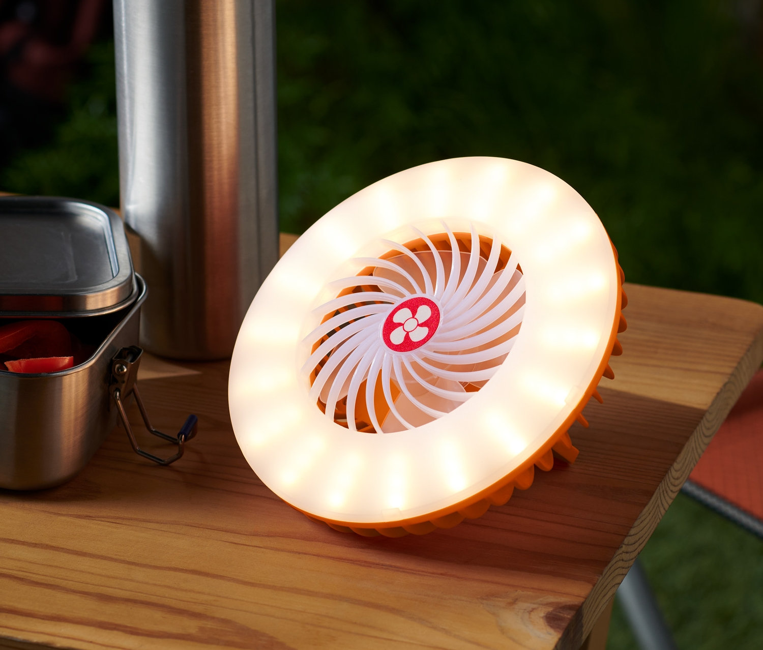 Na stole stojí LED ventilátor 2 v 1 vedle ocelové láhve a otevřeného jídlonosiče.
