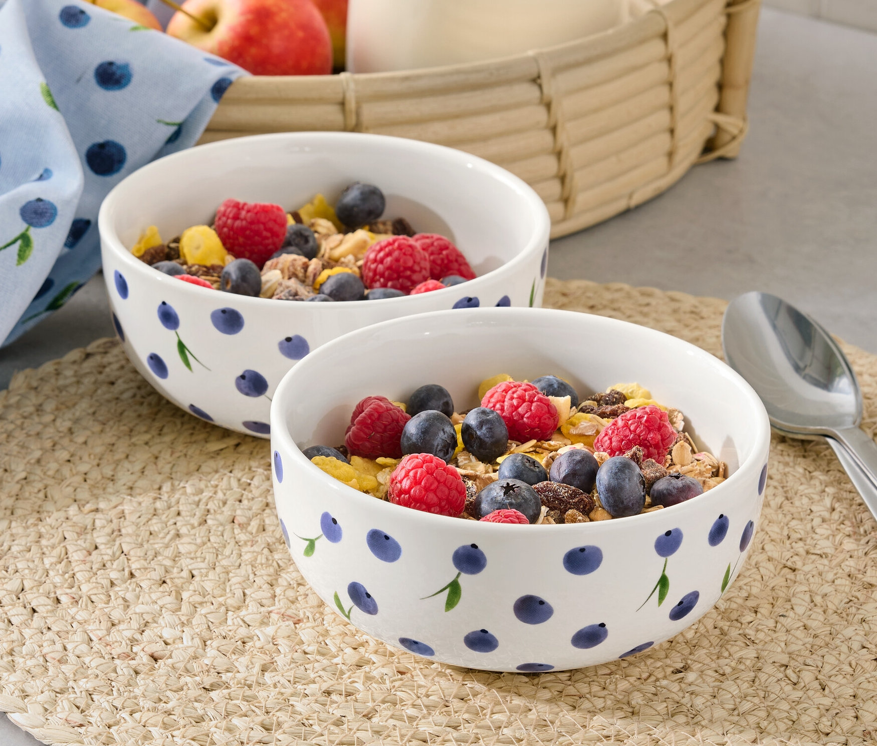 Dvě misky s müsli, malinami a borůvkami na prostírání. V pozadí džbán s mlékem a jablka v košíku.