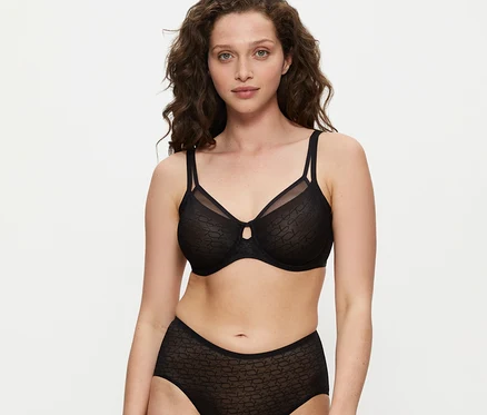 Žena s kudrnatými hnědými vlasy má na sobě černou zmenšující podprsenku Triumph Signature Sheer a ladící kalhotky s vysokým pasem, dívá se do kamery.