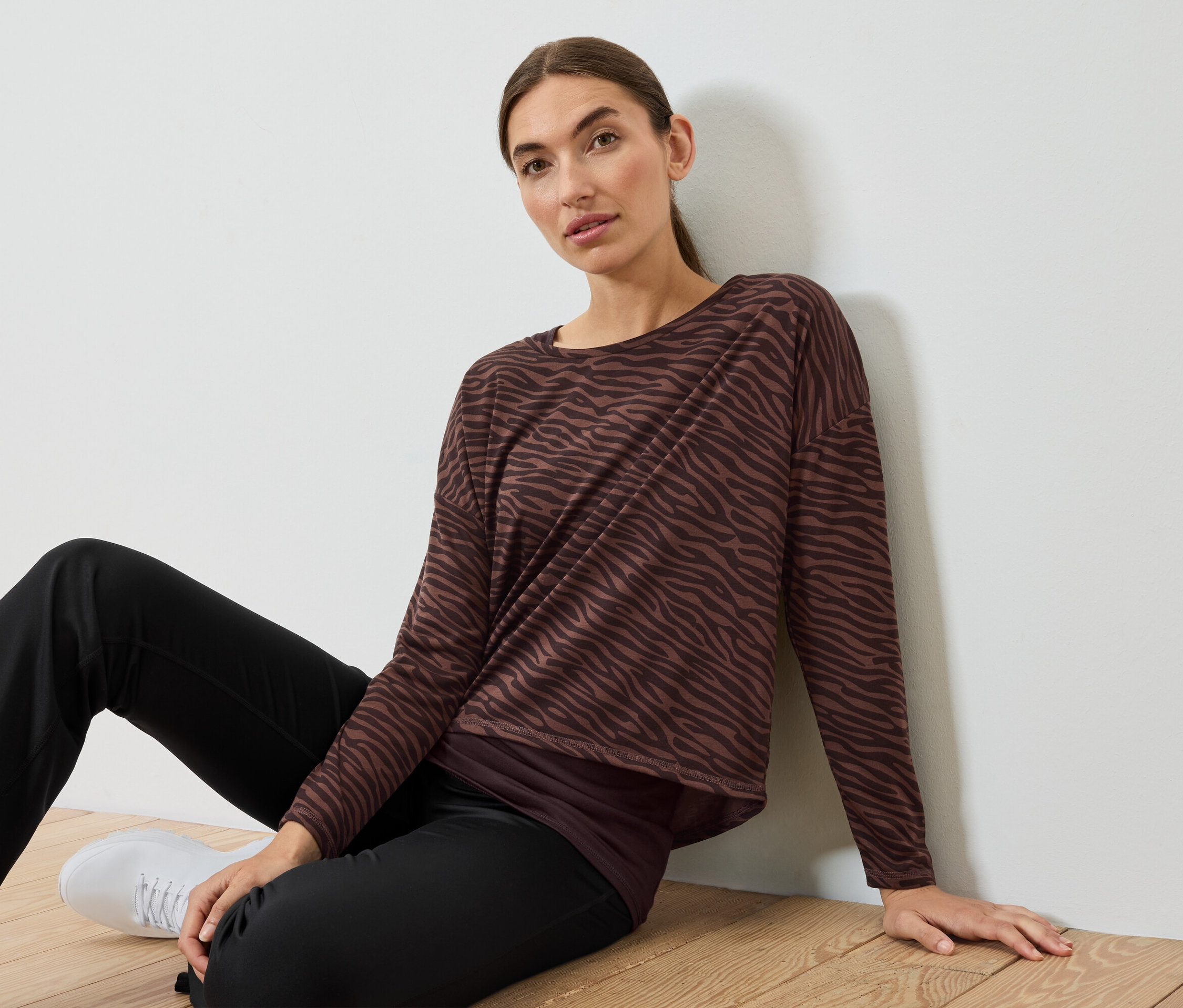 Žena sedí na podlaze v hnědo-černém zebrovaném topu a černých kalhotách.