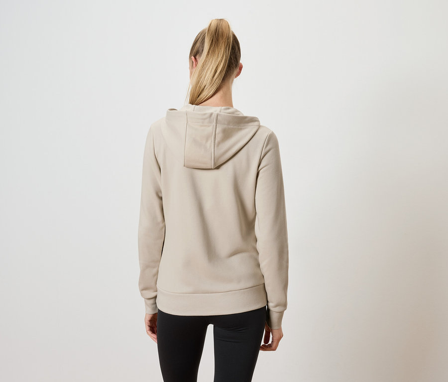 Zezadu je vidět žena s blond vlasy staženými do culíku, která má na sobě béžovou mikinu s kapucí a černé kalhoty.