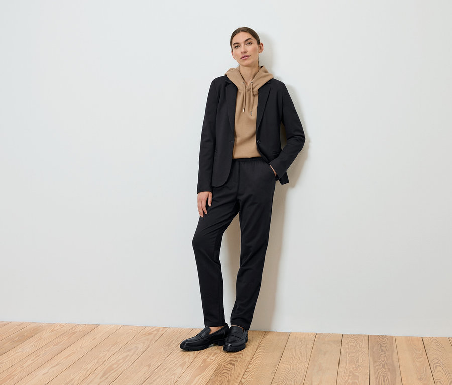 Žena pózuje na sobě černé sako, hnědou mikinu s kapucí a černé kalhoty.
