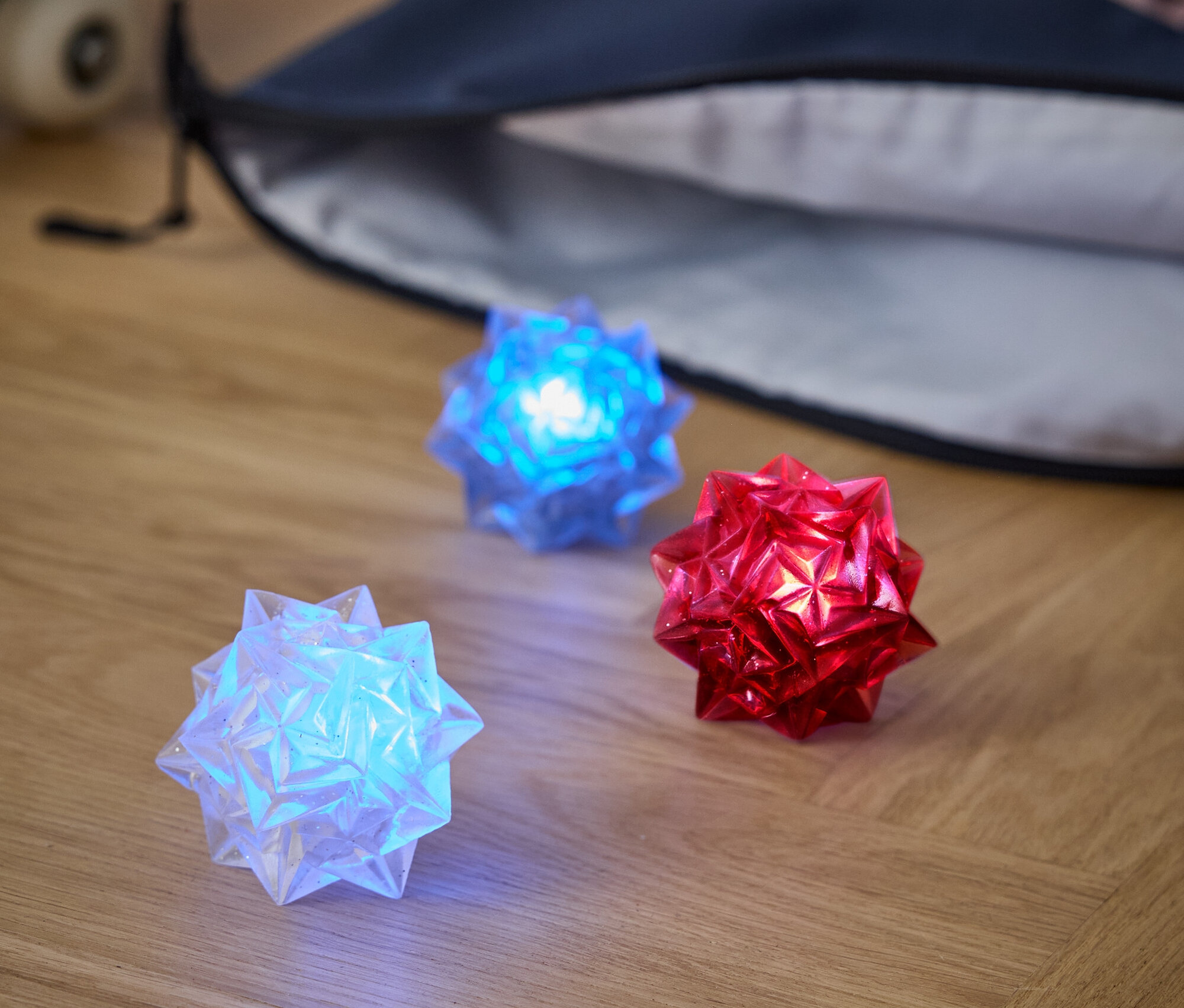 Na dřevěné podlaze leží tři světelné koule: bílá, modrá a červená. Za nimi je batoh a skateboard.