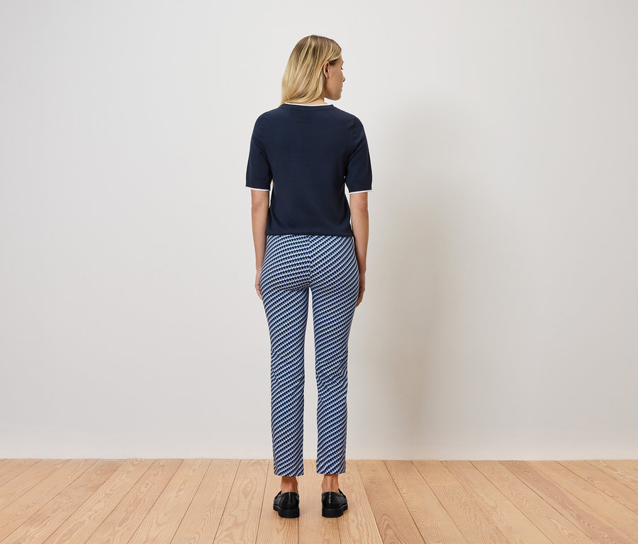 Blondýna stojí zády k fotoaparátu, má na sobě tmavě modrý top s krátkým rukávem a modrobílé pruhované kalhoty. Stojí na dřevěné podlaze.