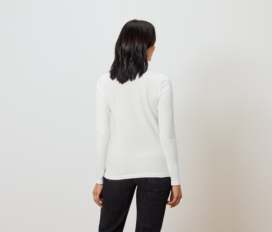 Žena s krátkými tmavými vlasy stojí zády a má na sobě bílou košili s dlouhým rukávem a černé džíny.