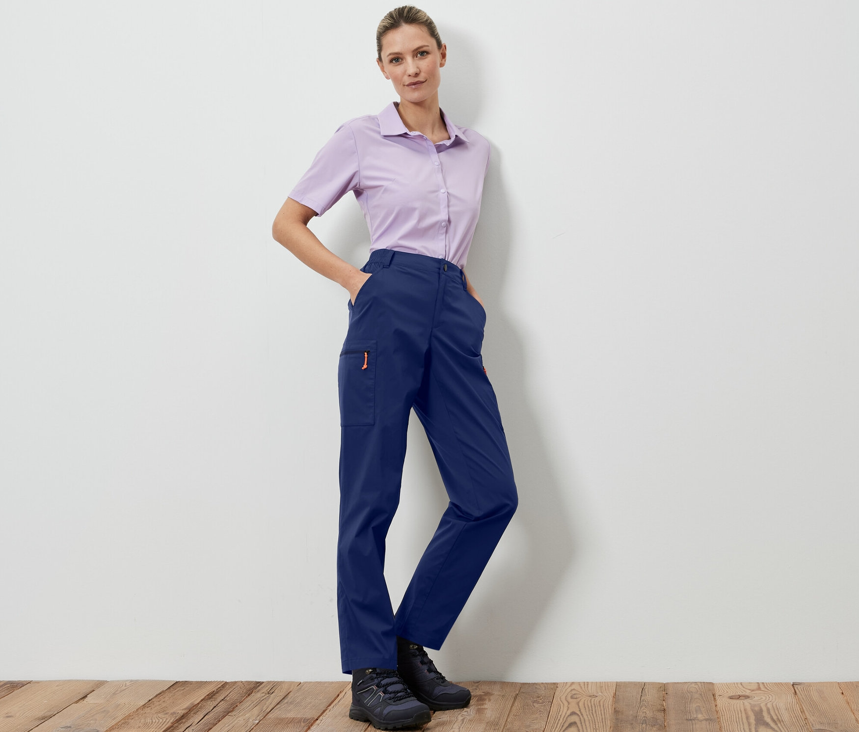 Žena pózuje v tmavě modrých sbalitelných outdoorových kalhotách a světle fialové košili.