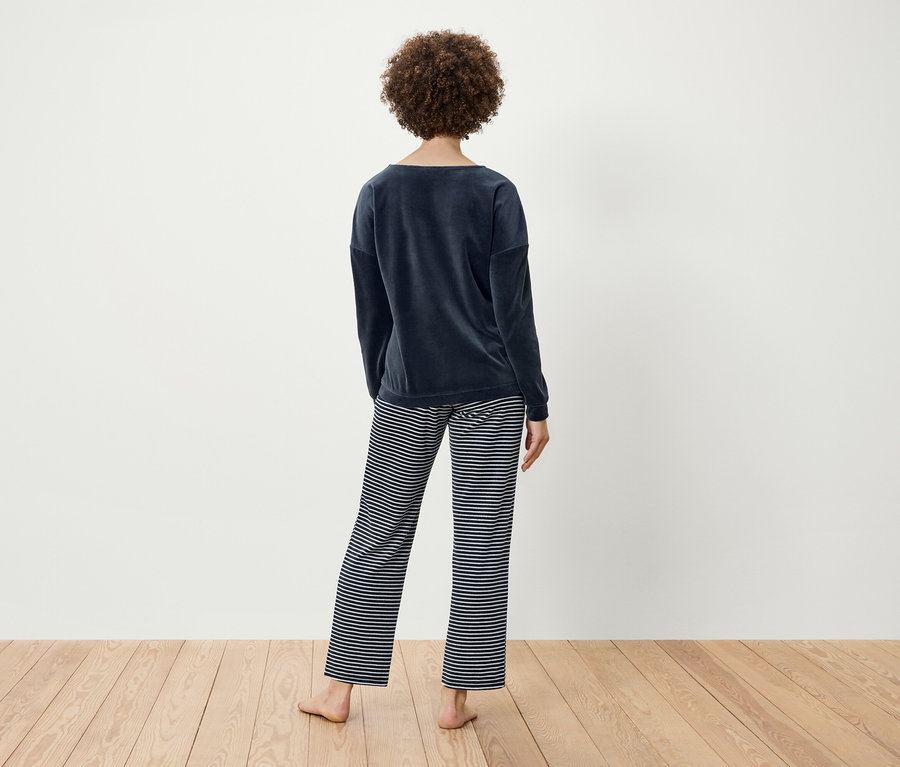 Žena s kudrnatými vlasy stojí zády a má na sobě tmavě modrý sametový top a pruhované kalhoty.