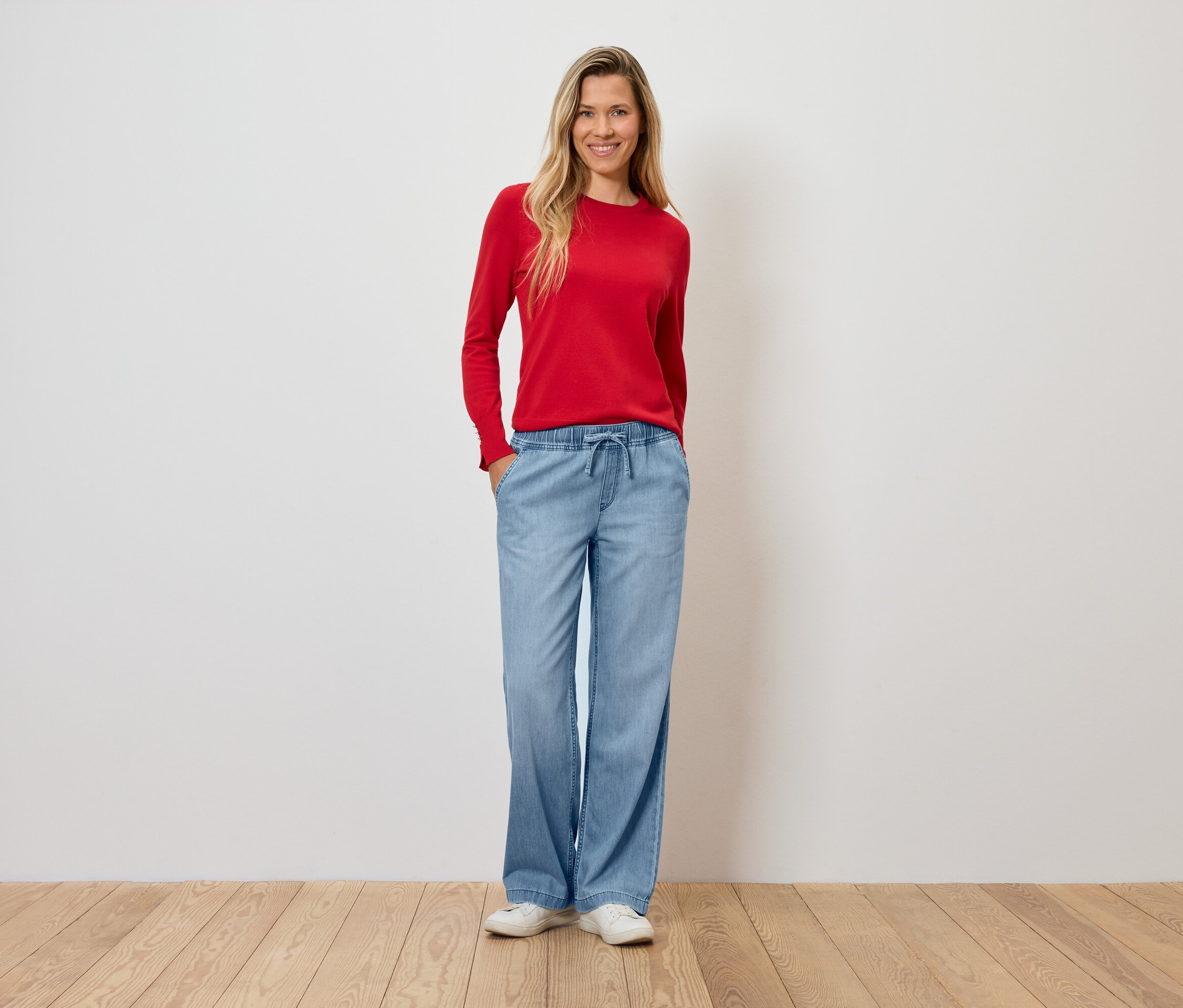 Žena v červeném svetru a širokých světle modrých kalhotách stojí na dřevěné podlaze.