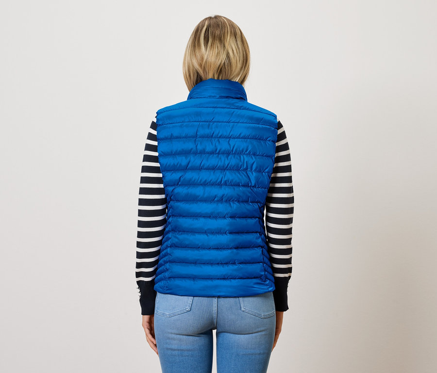 Žena zády k fotoaparátu má na sobě modrou prošívanou vestu a pruhovaný svetr.