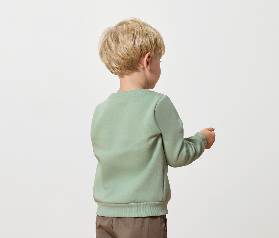 Chlapec s blond vlasy, oblečený v zelené mikině a hnědých kalhotách, stojí zády k fotoaparátu.